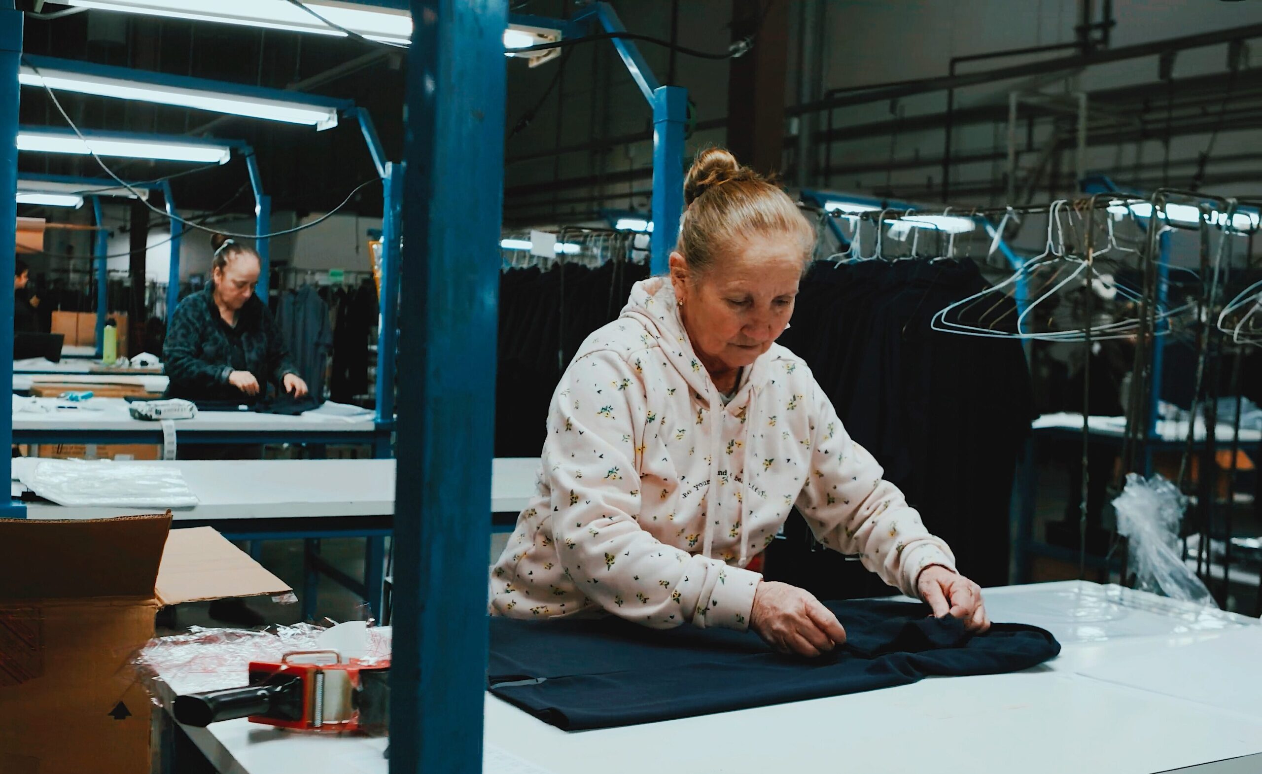 Wide view of the QCI floor: workers inspecting and folding garments at long blue worktables under strip lights
